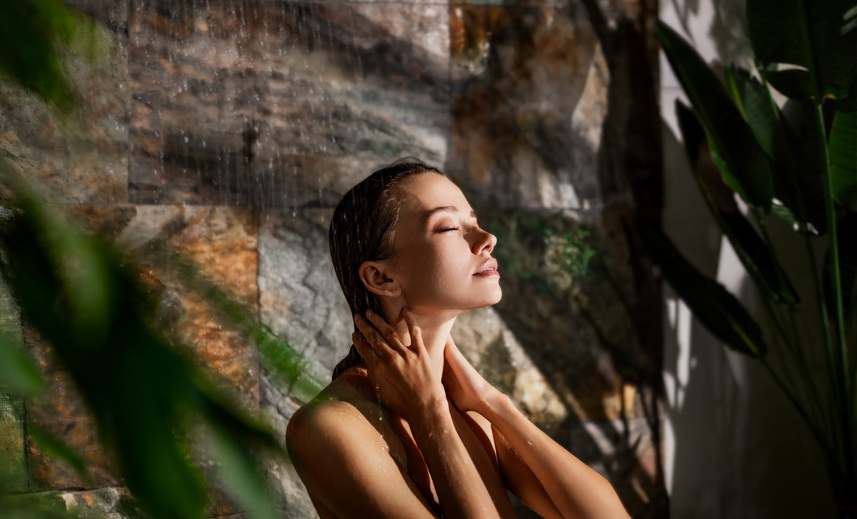 A woman standing in a sunlit shower with her eyes closed, one hand resting on her neck, warm light and greenery in the foreground.